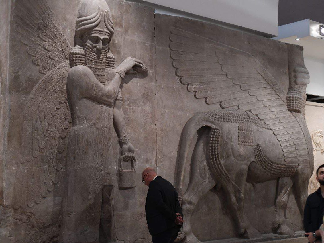 An Iraqi man is looking down in front of the sculpture of an Assyrian god